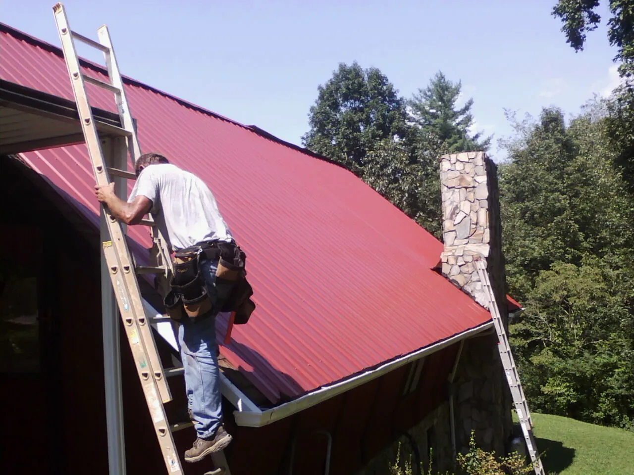 Roofer on a ladder beside a red metal roof and stone chimney — Soffit & Fascia Repair by Roofing Brothers in Zeeland